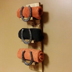 May include: A wooden towel rack with three shelves and three horseshoe towel holders. The towel rack is mounted on a wall and has three towels on it, one orange, one black, and one orange.