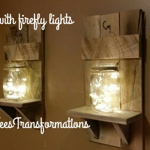 May include: Two rustic wooden shelves with a mason jar on each shelf. The jars are filled with fairy lights. The shelves are made of reclaimed wood and have a natural finish. The text "with firefly lights" and "Tees Transformations" is visible in the image.