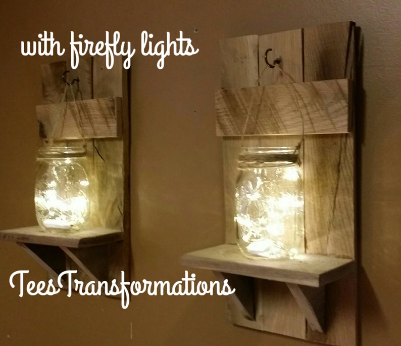 May include: Two rustic wooden shelves with a mason jar on each shelf. The jars are filled with fairy lights. The shelves are made of reclaimed wood and have a natural finish. The text "with firefly lights" and "Tees Transformations" is visible in the image.
