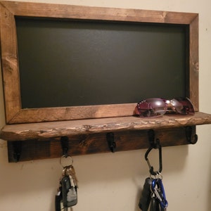 May include: A rustic wooden shelf with a chalkboard frame and hooks for hanging keys. The shelf is stained a dark brown and has a distressed finish. There are sunglasses on the shelf and keys hanging from the hooks.