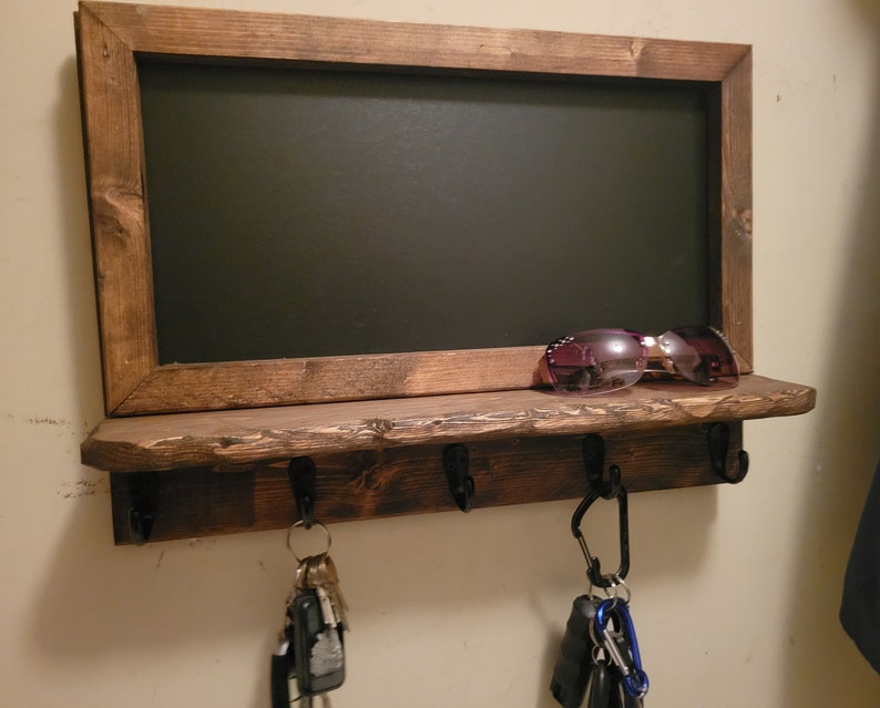 May include: A rustic wooden shelf with a chalkboard frame and hooks for hanging keys. The shelf is stained a dark brown and has a distressed finish. There are sunglasses on the shelf and keys hanging from the hooks.