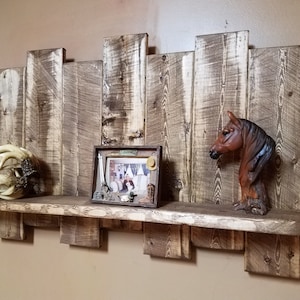 May include: Rustic wooden shelf with a distressed finish. The shelf has a decorative back made of reclaimed wood planks. The shelf is decorated with a small framed picture, a decorative horse head figurine, and a white decorative ball.