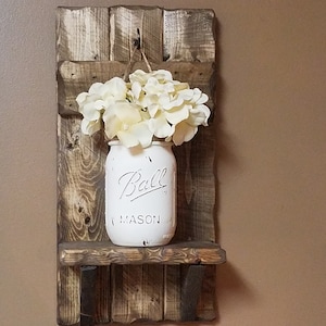 May include: A rustic wooden shelf with a white painted mason jar hanging from a hook. The jar is filled with white flowers and has the text "Ball Mason" on the front.