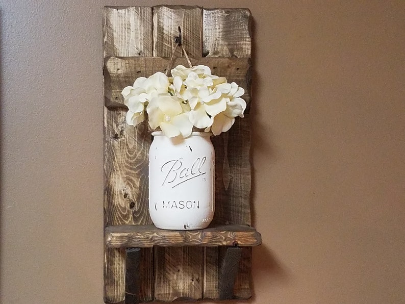 May include: A rustic wooden shelf with a white painted mason jar hanging from a hook. The jar is filled with white flowers and has the text "Ball Mason" on the front.