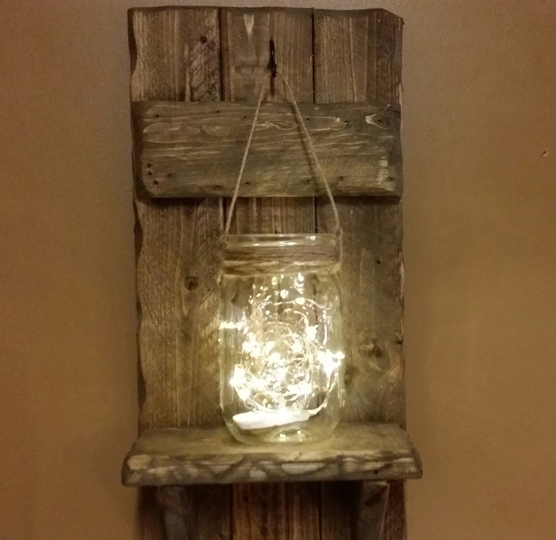 May include: A rustic wooden shelf with a hanging mason jar filled with fairy lights. The shelf is made of reclaimed wood and has a distressed finish.