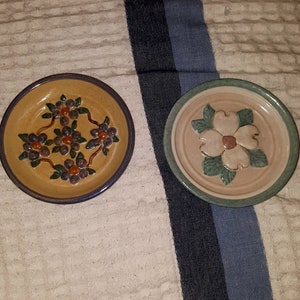 May include: Two ceramic plates with floral designs. One plate has a brown background with green leaves and purple flowers. The other plate has a beige background with green leaves and a white flower with a pink center.