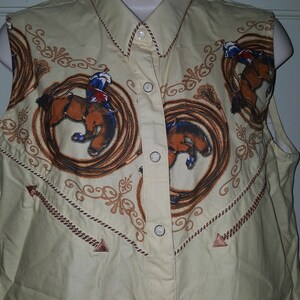May include: A sleeveless, light yellow button-down shirt with a western-style design. The shirt features a repeating pattern of cowboys riding horses in a lasso circle. The shirt has a pointed collar and brown trim.