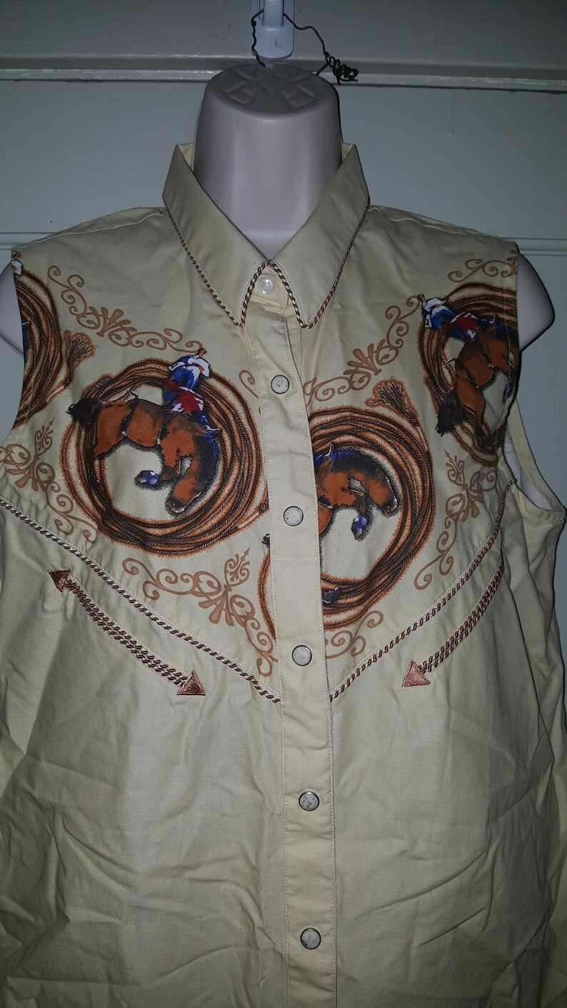 May include: A sleeveless, light yellow button-down shirt with a western-style design. The shirt features a repeating pattern of cowboys riding horses in a lasso circle. The shirt has a pointed collar and brown trim.