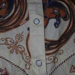 May include: A beige button-up shirt with a brown and white rope design and a brown and white horse design. The shirt has four white buttons down the front.