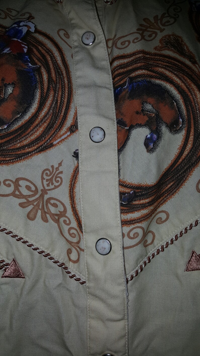May include: A beige button-up shirt with a brown and white rope design and a brown and white horse design. The shirt has four white buttons down the front.