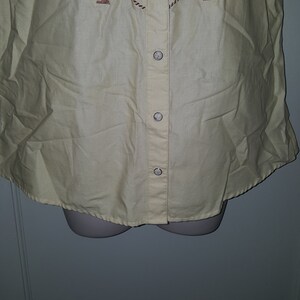May include: A sleeveless, light yellow button-up shirt with a brown and white print of a horse and rope design. The shirt has a button-up front and a pointed collar.