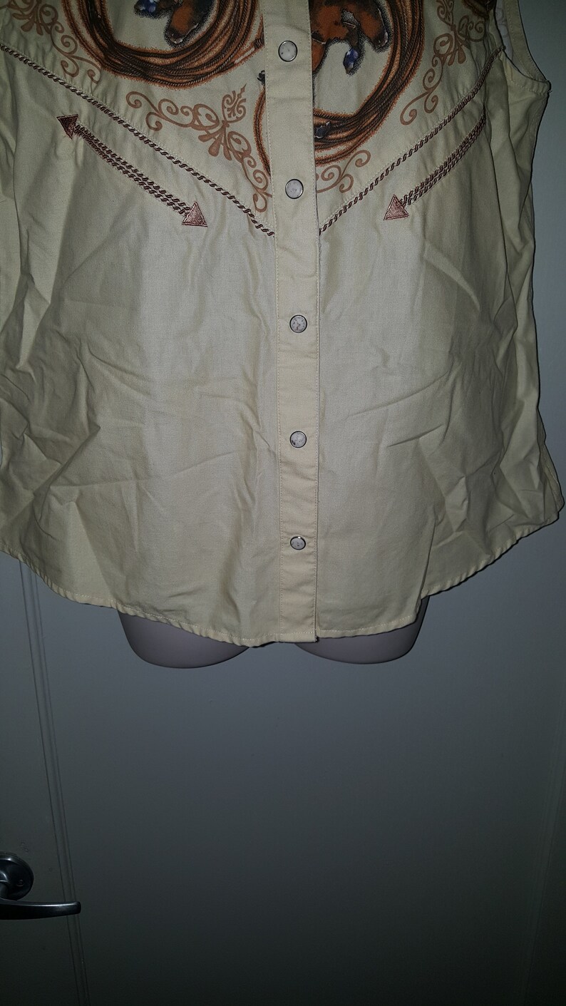 May include: A sleeveless, light yellow button-up shirt with a brown and white print of a horse and rope design. The shirt has a button-up front and a pointed collar.