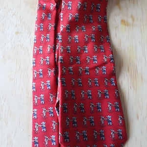 May include: A red silk tie with a repeating pattern of elephants wearing blue and white hats. The elephants are holding a red, white, and blue flag.