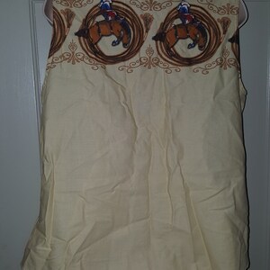 May include: A sleeveless, light yellow button-down shirt with a brown and white pattern of cowboys riding horses. The pattern is repeated across the top of the shirt.
