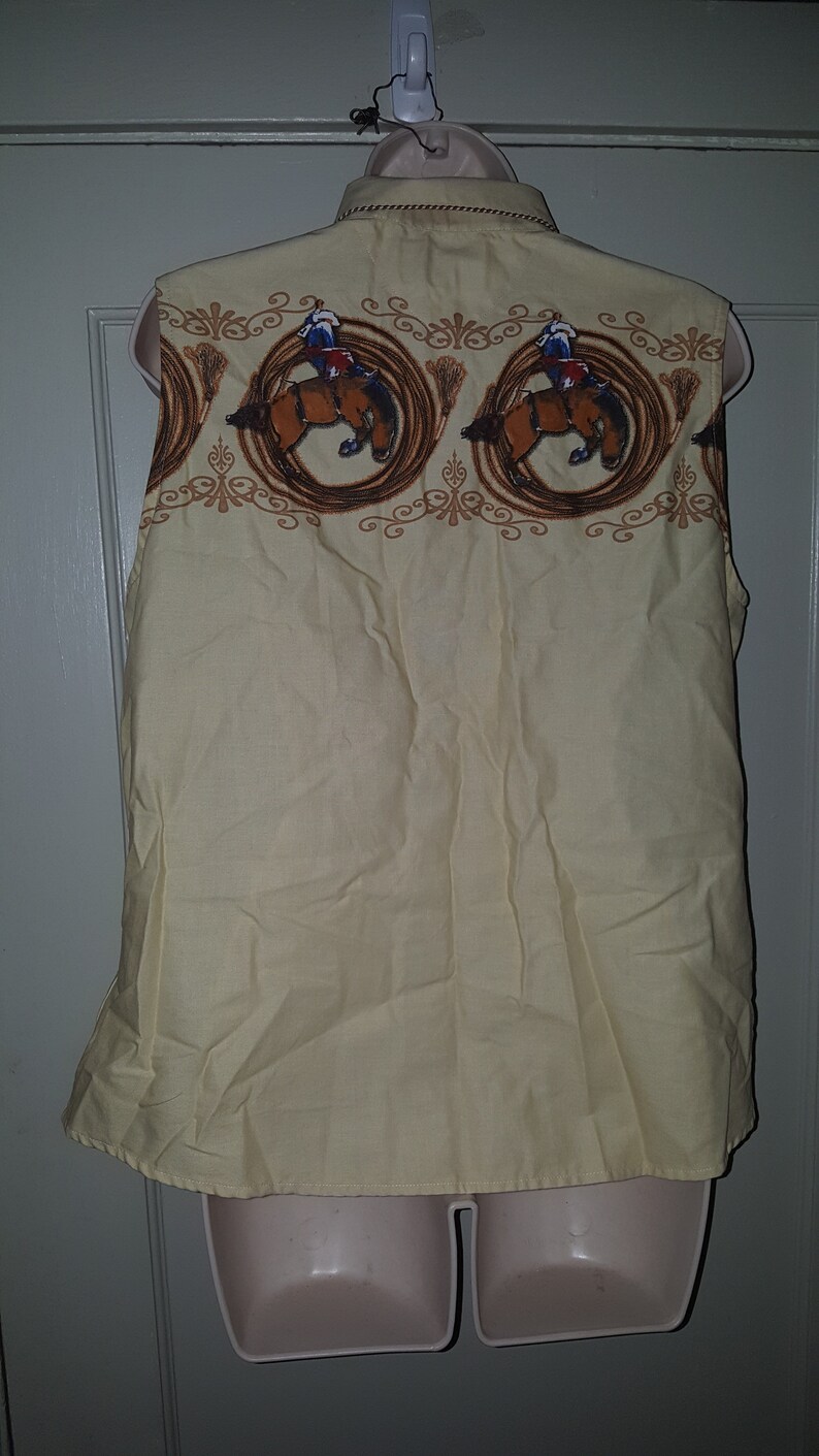 May include: A sleeveless, light yellow button-down shirt with a brown and white pattern of cowboys riding horses. The pattern is repeated across the top of the shirt.