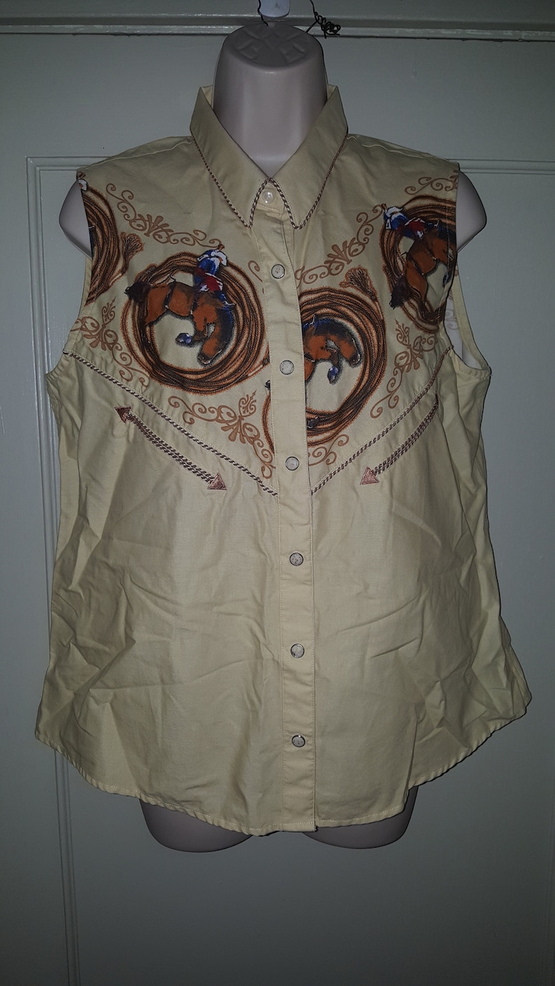 May include: A sleeveless, light yellow button-down shirt with a western-themed print featuring brown horses and rope designs. The shirt has a pointed collar and a button-up front.