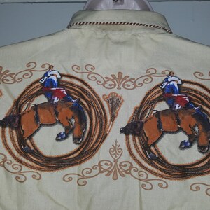 May include: A sleeveless, light beige button-down shirt with a repeating pattern of cowboys riding bucking horses. The horses are brown and the cowboys are wearing blue and red clothing. The pattern is surrounded by a brown rope design.