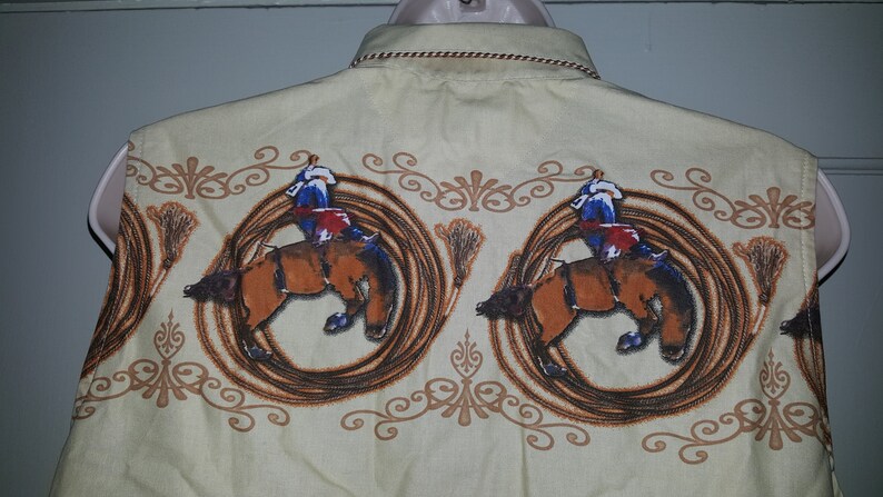 May include: A sleeveless, light beige button-down shirt with a repeating pattern of cowboys riding bucking horses. The horses are brown and the cowboys are wearing blue and red clothing. The pattern is surrounded by a brown rope design.