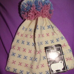May include: White knitted beanie with a pink, blue, and white pom pom on top. The beanie has a pink and blue geometric pattern. A black tag with white text reads "The Alpen".