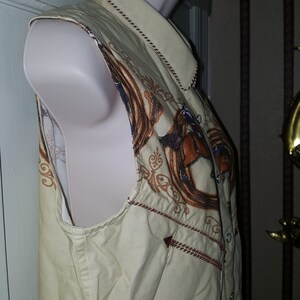 May include: A sleeveless, beige button-up shirt with a brown and gold embroidered design of a horse and a horseshoe. The shirt has a brown trim around the collar and armholes.