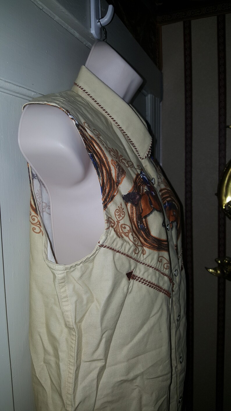 May include: A sleeveless, beige button-up shirt with a brown and gold embroidered design of a horse and a horseshoe. The shirt has a brown trim around the collar and armholes.