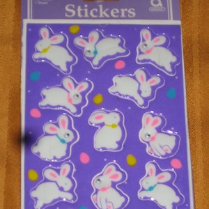 May include: A sheet of stickers featuring 10 white bunny rabbits with pink ears and blue bows. The stickers are on a purple background with yellow, pink, and blue dots.