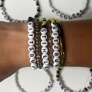 May include: A set of seven beaded bracelets with black and white lettering. The bracelets spell out "The Burgh", "Pittsburgh", "Stillers", "Blitzburgh", "Steelers", "Game Day", and "Black and Gold".