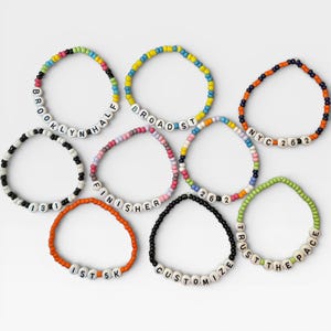 May include: A collection of beaded bracelets in various colors. Each bracelet features white letter beads spelling out words like "BROOKLYN HALF", "BROAD ST", "NYC 262", "FINISHER", "1ST 5K", and "CUSTOMIZE".