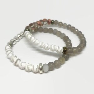 May include: Two bracelets made of gray and white agate beads. One bracelet has a white and gray swirl pattern, and the other has solid gray beads. The bracelets are stacked on top of each other.