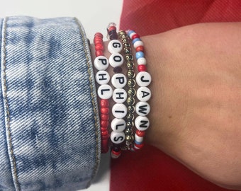 May include: A stack of beaded bracelets with the words "GO PHILS" and "JAWN" spelled out in black letters on white beads. The bracelets are red, white, and blue.