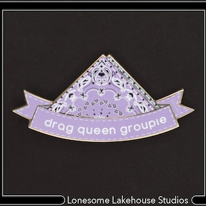 May include: Purple enamel pin featuring a bandana design and the text "drag queen groupie" in white lettering.