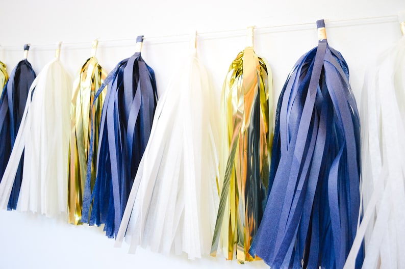May include: A garland of tissue paper tassels in navy blue, white, and gold. The tassels are strung together on a white string and are hanging from a white wall.