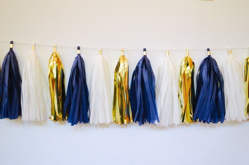 May include: A string of tissue paper tassels in navy blue, white, and gold. The tassels are strung together with a white string and are hanging from a white wall.