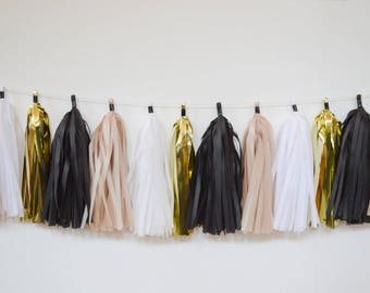 May include: A garland of tissue paper tassels in black, white, gold, and beige. The tassels are strung on a white string and are hanging from a white wall.