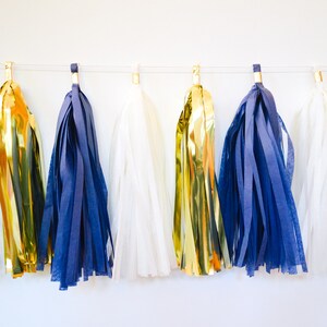 May include: A garland of tissue paper tassels in white, gold, and navy blue. The tassels are strung on a white string and are hanging from a white wall.