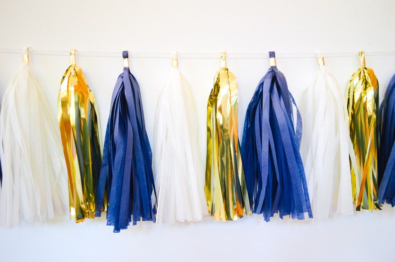 May include: A garland of tissue paper tassels in white, gold, and navy blue. The tassels are strung on a white string and are hanging from a white wall.