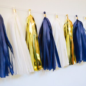 May include: A garland of tissue paper tassels in navy blue, white, and gold. The tassels are strung together with a white string.