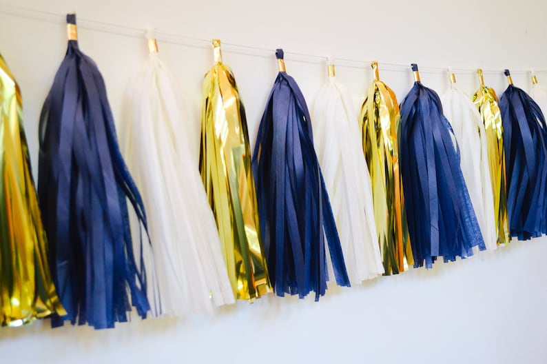 May include: A garland of tissue paper tassels in navy blue, white, and gold. The tassels are strung together with a white string.