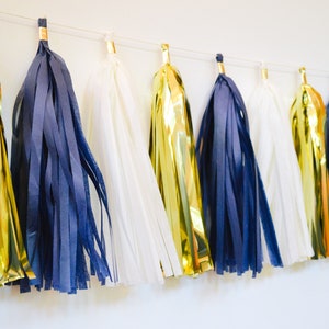 May include: A garland of tissue paper tassels in gold, navy blue, and white. The tassels are strung together with white string and are hanging from a white wall.