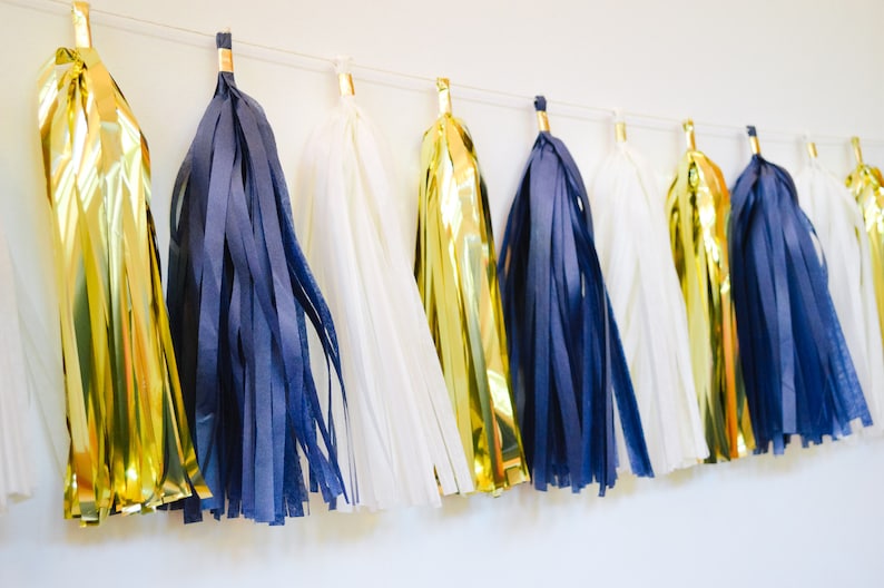 May include: A garland of tissue paper tassels in gold, navy blue, and white. The tassels are strung together with white string and are hanging from a white wall.