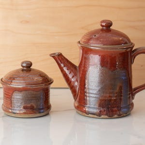 May include: A set of two ceramic pottery pieces in a reddish-brown glaze with a speckled pattern. The larger piece is a teapot with a lid and a handle. The smaller piece is a lidded jar.