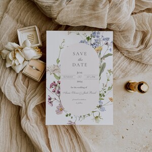 May include: Save the Date card with a floral design featuring a wreath of purple, blue, and yellow flowers. The text reads "Save the Date", "Sunday, June 20, 2024, at 5 PM", "for the wedding of Anne Oliver & Jack Bond", and "formal invitations to follow".