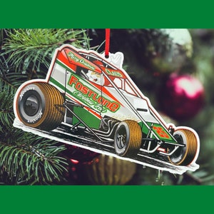 May include: A white, green, and red race car ornament with the words "Fastlane Christmas Tree" and "Merry Christmas" on it. Santa Claus is driving the car.