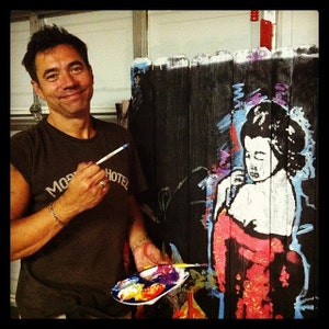 May include: A man wearing a brown t-shirt with the text "More Hotel" painted on it, is painting a mural of a woman in a red kimono on a black wooden fence. He is holding a paintbrush and a palette of paint.