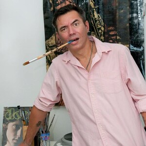 Puede incluir: Un hombre con una camisa de botones rosa y pantalones de camuflaje sostiene un pincel en la boca. Est&aacute; de pie frente a una pared de madera con una pintura en ella. La pintura es de una persona tocando el saxof&oacute;n. Hay pinceles en una lata sobre una mesa blanca frente a &eacute;l.