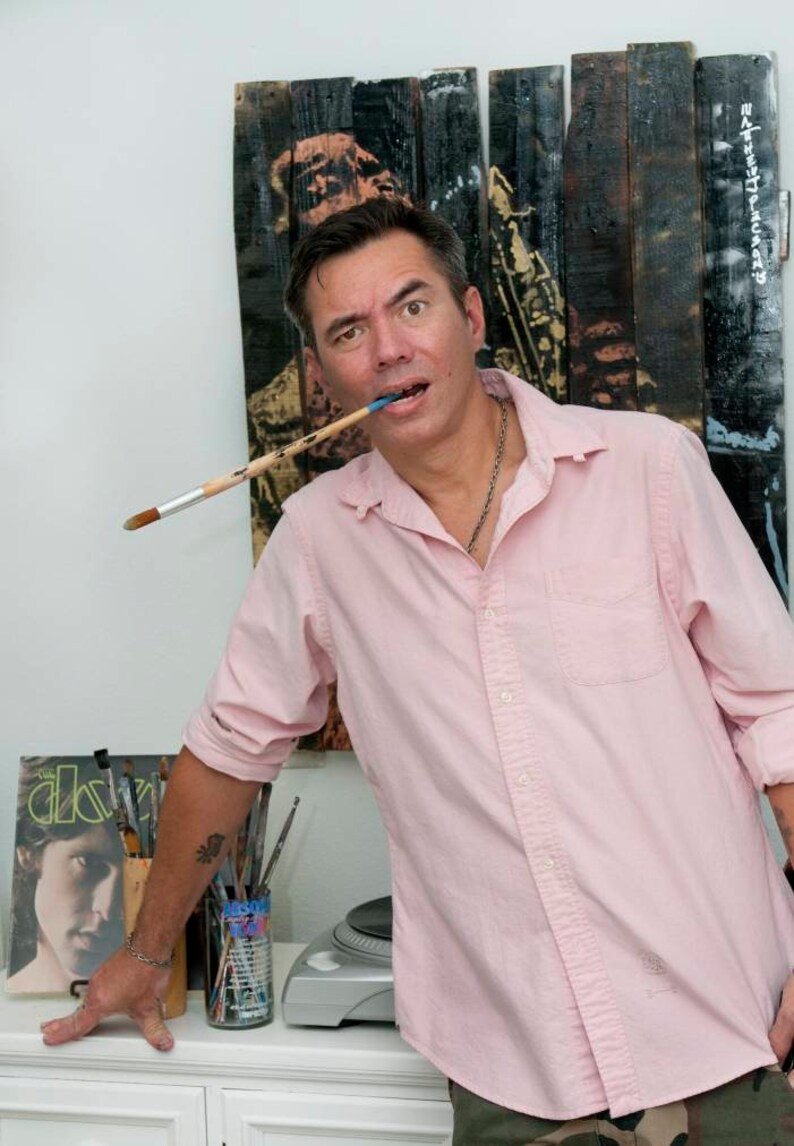 Puede incluir: Un hombre con una camisa de botones rosa y pantalones de camuflaje sostiene un pincel en la boca. Est&aacute; de pie frente a una pared de madera con una pintura en ella. La pintura es de una persona tocando el saxof&oacute;n. Hay pinceles en una lata sobre una mesa blanca frente a &eacute;l.