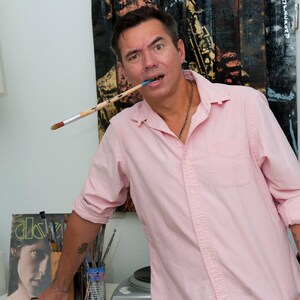 May include: A man wearing a pink button-down shirt and camouflage pants holds a paintbrush in his mouth. He is standing in front of a wooden pallet art piece with a figure playing a saxophone. The man has a tattoo on his arm.