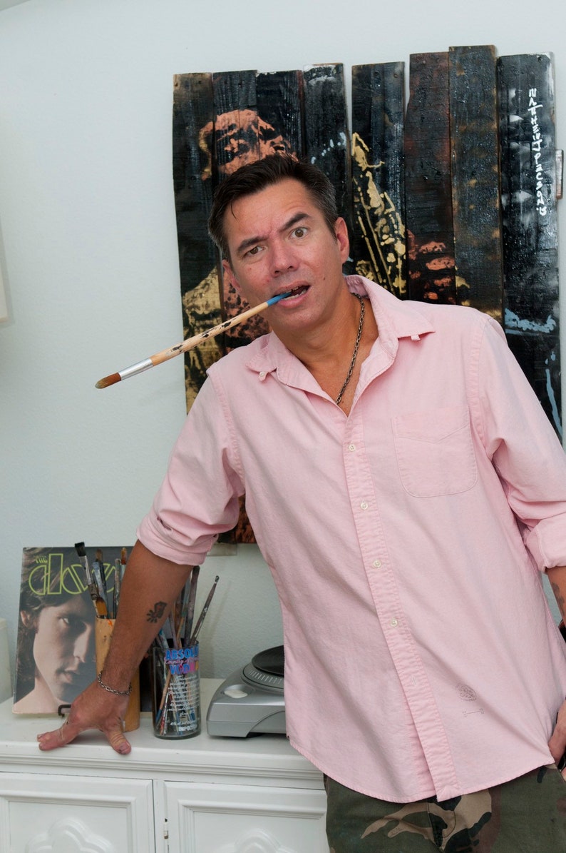 May include: A man wearing a pink button-down shirt and camouflage pants holds a paintbrush in his mouth. He is standing in front of a wooden pallet art piece with a figure playing a saxophone. The man has a tattoo on his arm.