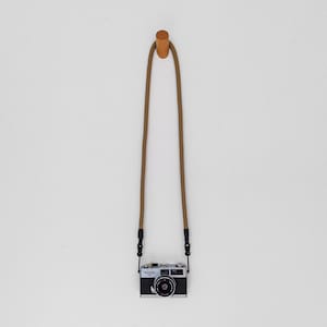 May include: A vintage silver and black camera with a brown strap hangs from a wooden peg on a white wall. The camera has a lens and several dials. The strap is attached to the camera with black clips.