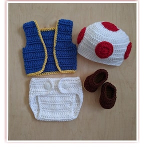 May include: A crocheted baby outfit in blue, white, and red. The outfit includes a vest, diaper cover, hat, and shoes. The hat is shaped like a mushroom.